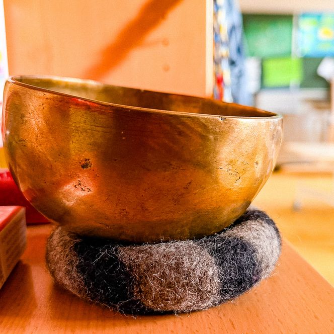 Eine goldfarbene Klangschale auf einem Filzuntersetzer im Vordergrund, mit dem hellen Gruppenraum der nachschulischen Betreuung Soderstorf im unscharfen Hintergrund.