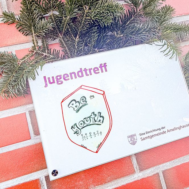 Ein Schild mit der Aufschrift „Jugendtreff Be-Youth“ hängt an einer roten Backsteinwand, geschmückt mit Tannenzweigen.