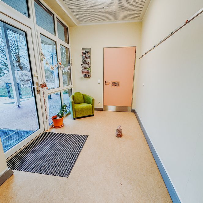 Heller Eingangsbereich des Kindergartens Rehlingen mit großen Fenstern, einer grünen Sitzgelegenheit, einer Pflanze im Blumentopf und einer hellrosa Tür.