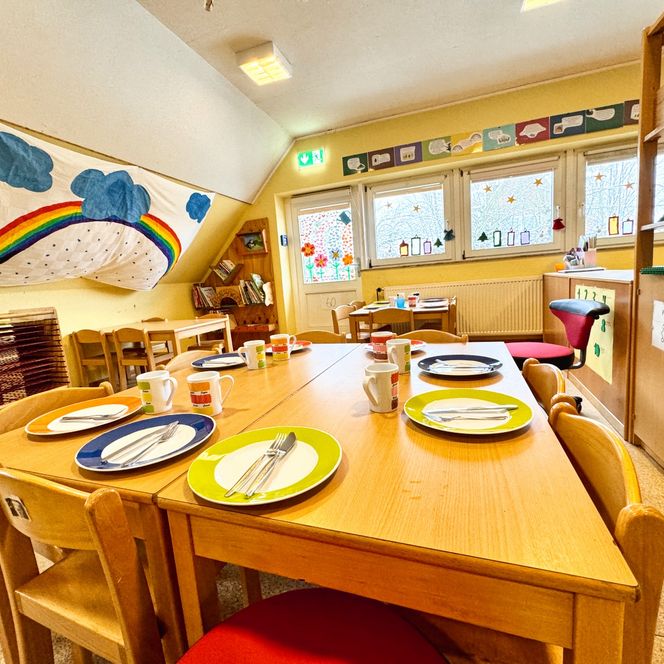 Ein Speiseraum im Kindergarten mit gedeckten Holztischen, bunten Tellern und Tassen sowie dekorierten Fenstern im Hintergrund.