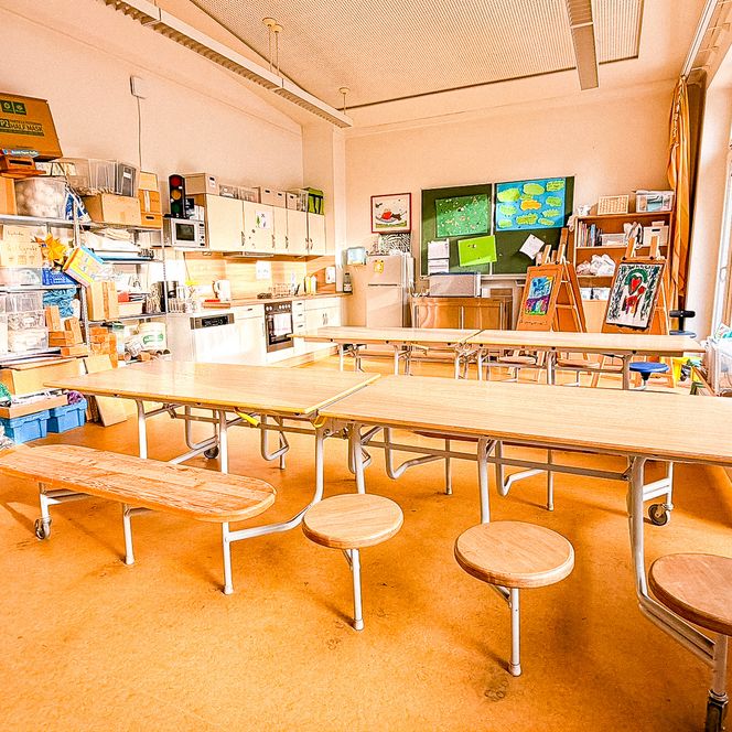Ein heller Gruppenraum mit langen Holztischen, integrierten Sitzplätzen und Bastelmaterialien an den Wänden.