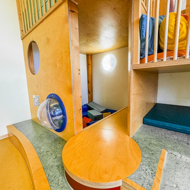 Spiel- und Kletterbereich in der Kinderkrippe Amelinghausen mit Holzkonstruktion, Kissen, Matten und Durchguck-Elementen.