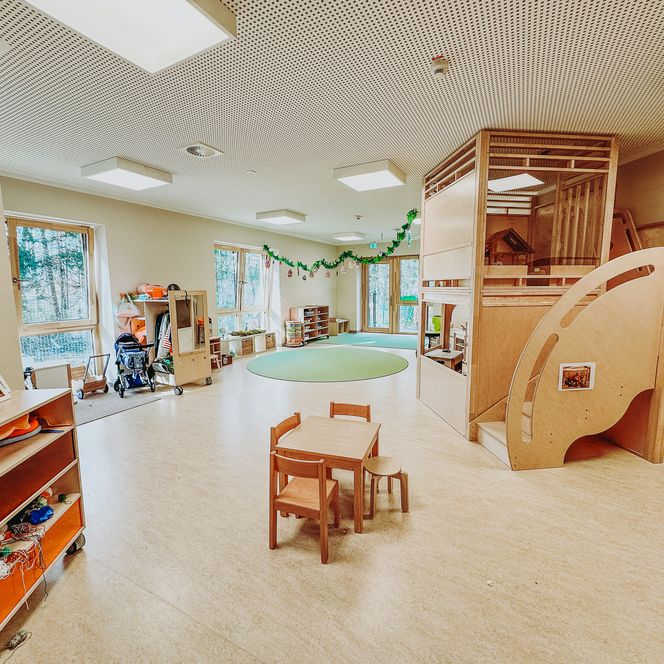 Heller Gruppenraum in der KiTa Soderstorf mit Holzmöbeln, einem runden grünen Teppich und einer großen Spielebene aus Holz.