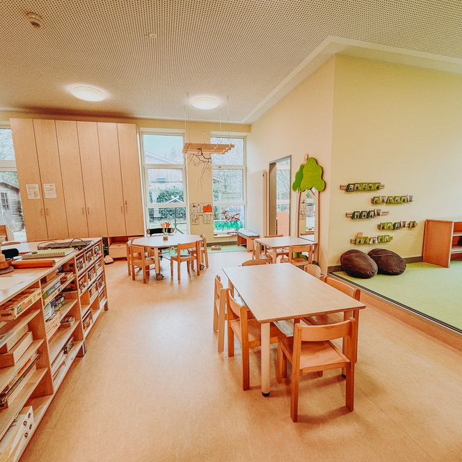 Heller Gruppenraum im Kindergarten Rehlingen mit Tischen, Stühlen, offenen Regalen, Spielen und angrenzender Spielecke mit grünem Teppich.