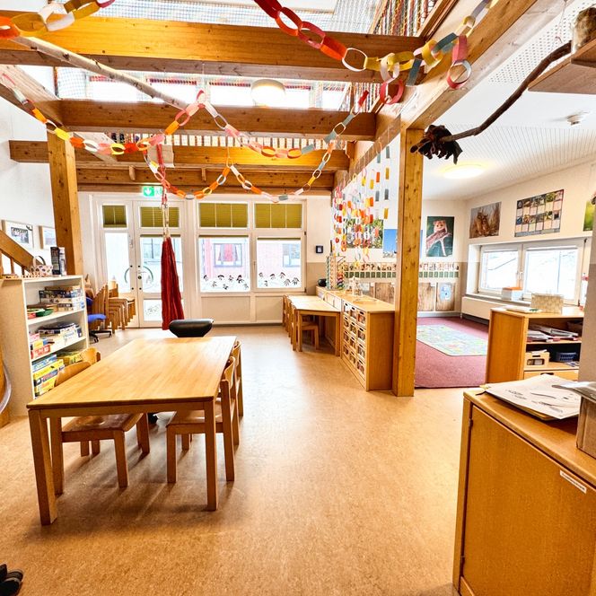 Ein großer Gruppenraum im Kindergarten mit Holztischen, Regalen mit Spielen, bunter Wanddekoration und einer Spielecke mit Teppich.