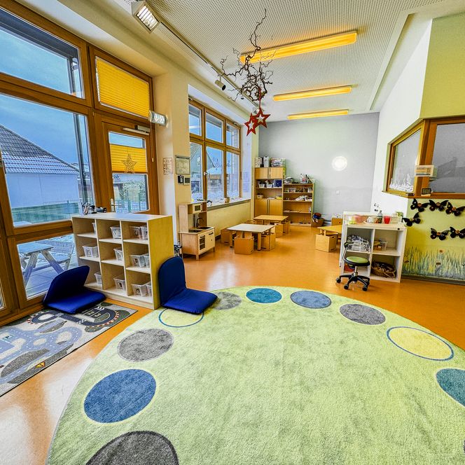 Heller Gruppenraum in der Kinderkrippe Amelinghausen mit großem Teppich, Spielbereichen, Tischen und großen Fenstern.