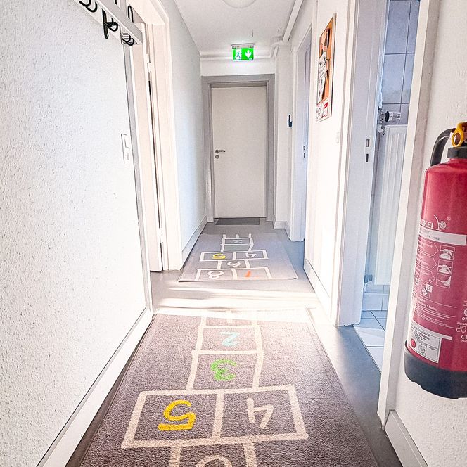 Ein heller, weißer Flur im Jugendtreff „Be-youth“ mit einem Teppichläufer im „Himmel-und-Hölle“-Hüpfspiel-Design und einem Feuerlöscher im Vordergrund.