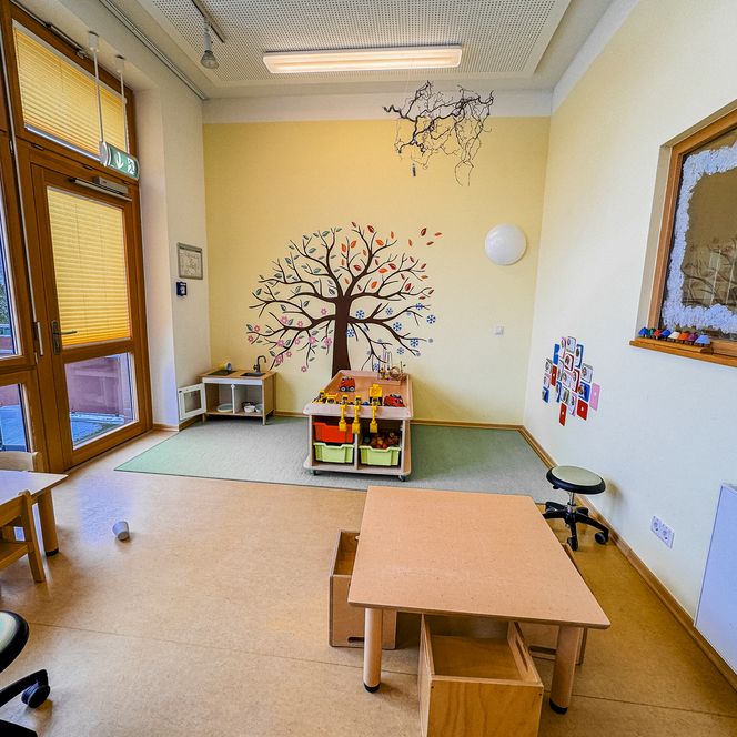 Spielbereich in der Kinderkrippe Amelinghausen mit kleinen Holztischen, Stühlen und Wandgestaltung mit buntem Lebensbaum.