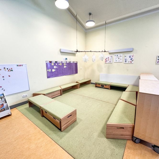 Ein Gruppenraum im Kindergarten mit grünen Sitzpodesten, Magnettafel, Bücherregal und Wanddekoration mit Kinderzeichnungen.