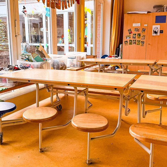 Eine helle Sitzgruppe mit Holztischen und runden Hockern in der nachschulischen Betreuung Soderstorf, direkt vor einer großen Fensterfront mit Blick in den Garten.