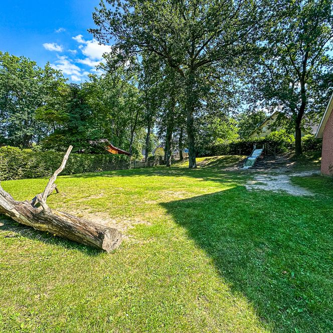 Grüne Wiese am Kindergarten Oldendorf/Luhe mit einem großen Baumstamm zum Balancieren und einer Rutsche am Hang.
