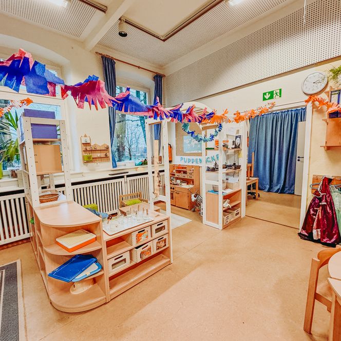 Gruppenraum in der Kita Betzendorf mit Puppenecke, Regalen voller Spielmaterialien, runden Tischen und bunter Papierketten-Dekoration.