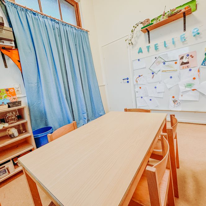 Ein Atelierraum im Kindergarten mit Holztisch, Stühlen, Regalen und einer Wand mit Kinderzeichnungen.