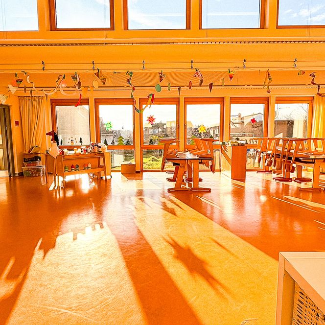 Ein sonnendurchfluteter Gruppenraum mit großen Fenstern, bunten Bastelarbeiten und Holztischen.