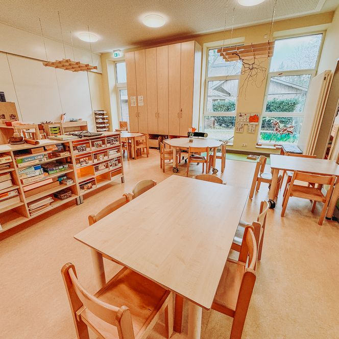 Heller Gruppenraum im Kindergarten Rehlingen mit mehreren Holztischen und Stühlen, Regalen voller Spiele und Bastelmaterial sowie großen Fenstern, die viel Tageslicht hineinlassen.