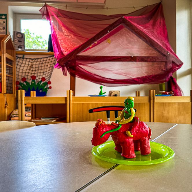 Eine bunte Spielfigur aus Knete: ein grünes Männchen reitet auf einem roten Tier, aufgestellt auf einem grünen Teller in einem Kindergartenraum.