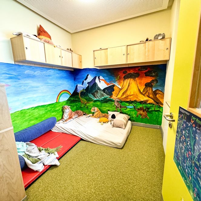 Ein Ruhe- und Kuschelraum im Kindergarten mit Matten, Kissen und einer bunten Wandbemalung mit Tieren, Bergen und Vulkanen.