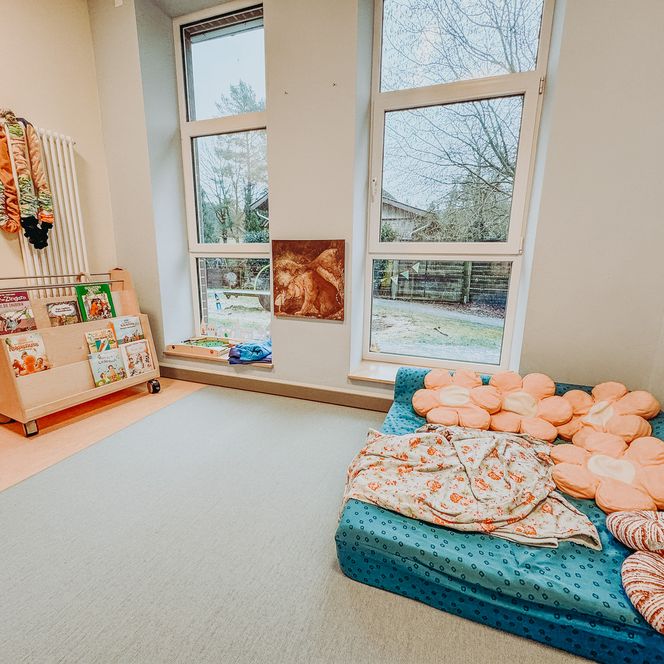Helle Leseecke im Kindergarten Rehlingen mit Sofa, Blumen-Sitzkissen, Decke und einem Bücherwagen mit bunten Kinderbüchern.
