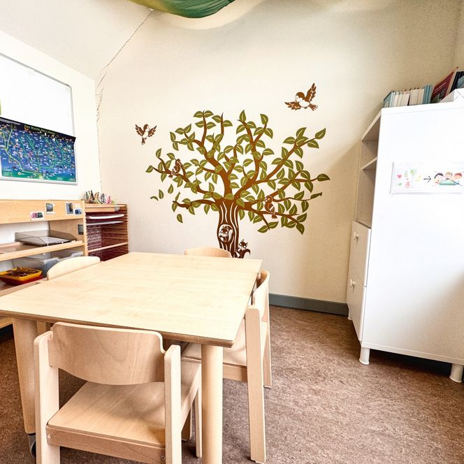 Ein heller Raum im Kindergarten mit Holztisch und Stühlen, Regalen, einem Schrank und einer Wandgestaltung mit Baum und Vögeln.