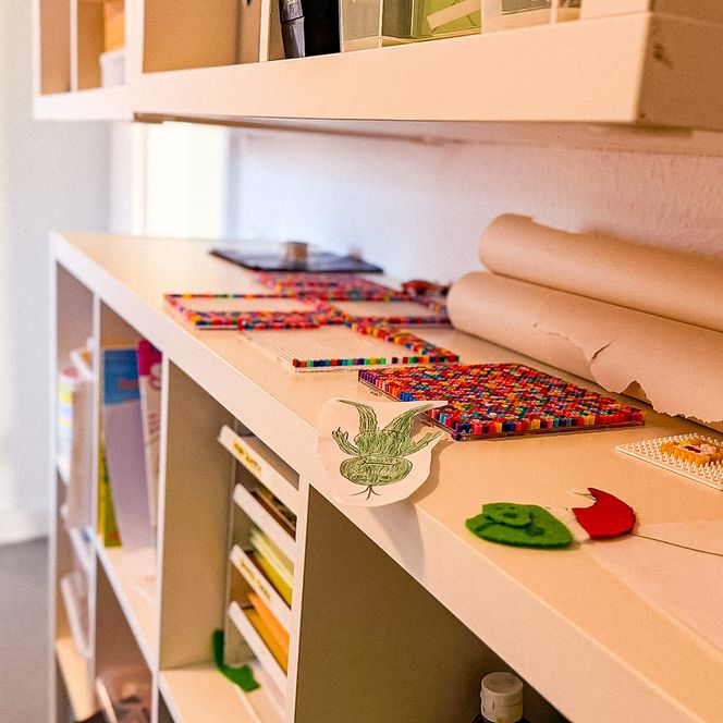 Nahaufnahme von Bastelmaterialien wie Bügelperlen, Papierrollen und Zeichnungen auf einem weißen Regal im Jugendtreff „Be-youth“.