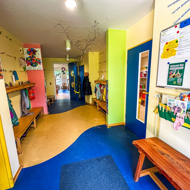 Bunter Flur im Kindergarten Oldendorf/Luhe mit Garderobenhaken, Sitzbänken, Elterninformationen an einer Pinnwand und geöffneter blauer Eingangstür im Hintergrund.