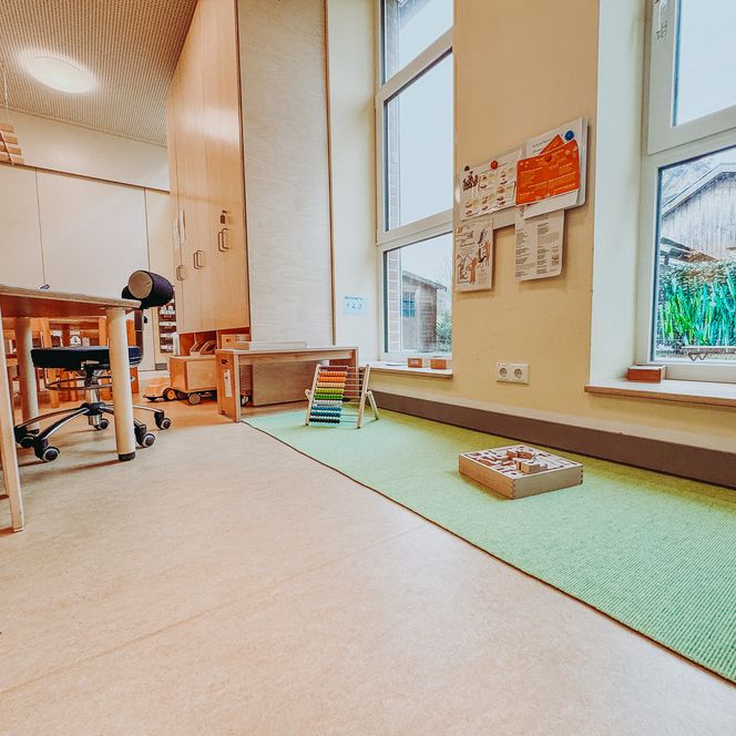Lern- und Spielecke im Kindergarten Rehlingen mit grünem Teppich, Motorik Spielzeug und großen Fenstern mit Tiermalerei.