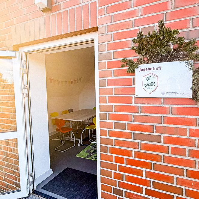 Blick durch die offene Tür in einen hellen Raum des Jugendtreffs „Be-youth“ mit bunten Stühlen und einem Tisch, neben dem Eingang hängt das offizielle Schild der Einrichtung.