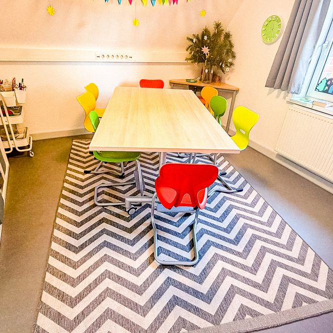 Ein heller Raum im Jugendtreff „Be-youth“ mit einem großen zentralen Tisch, bunten Stühlen, einem modernen Teppich mit Zickzack-Muster und weihnachtlicher Fensterdekoration.