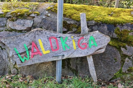 Ein handbemaltes Holzschild mit der Aufschrift „Waldkiga“ vor einer Steinmauer mit Moosbewuchs.
