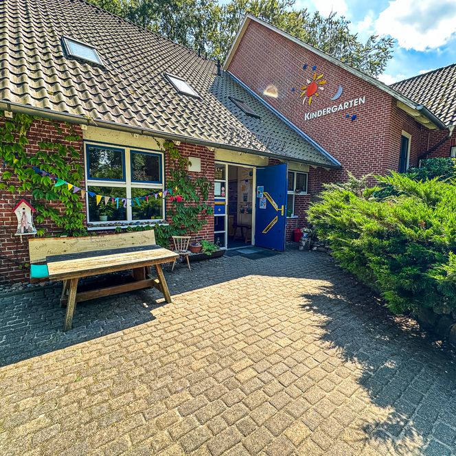 Eingangsbereich des Kindergartens Oldendorf/Luhe mit geöffneter blauer Tür, bepflanzter Fassade, bunter Wimpelkette und Holzbank davor.