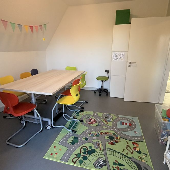 Innenansicht eines hellen Gruppenraums im Jugendtreff „Be-youth“ mit einem großen Arbeitstisch, bunten Stühlen, einer Wimpelkette und einem Spielteppich.