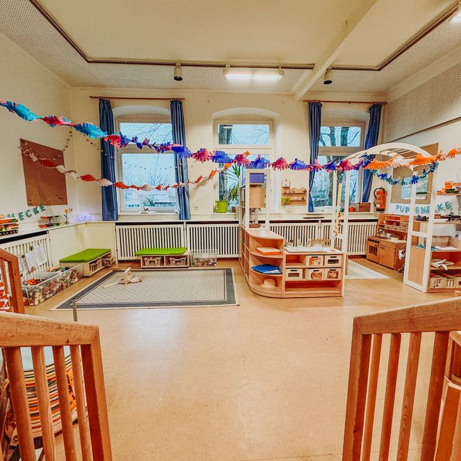 Ein Gruppenraum im Kindergarten mit bunter Girlandendekoration, Spielecke mit Regalen und einer Puppenecke.