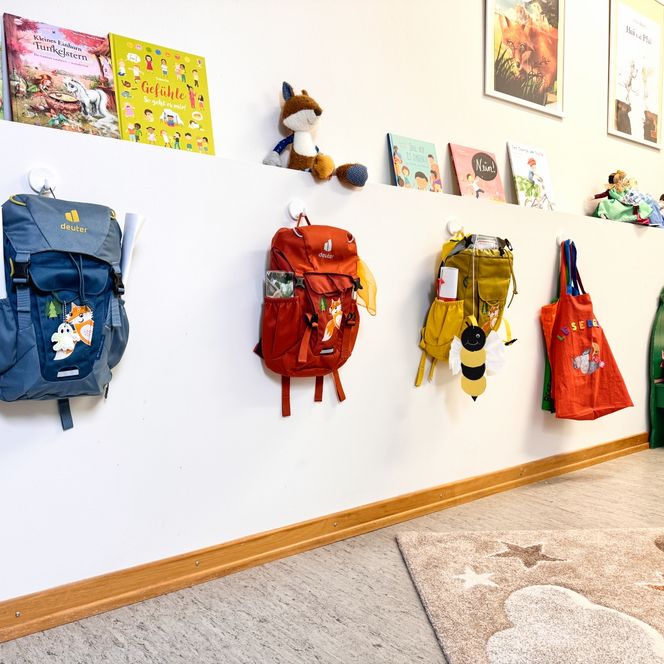 Mehrere bunte Kinderrucksäcke hängen im Kindergarten an Haken an der Wand, darüber stehen Kinderbücher und Spielzeug.