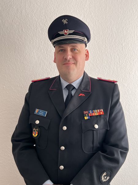 „Dennis Knoblich, Gemeindebrandmeister der Samtgemeinde Amelinghausen, steht in Uniform vor einer hellen Wand und blickt freundlich in die Kamera.“