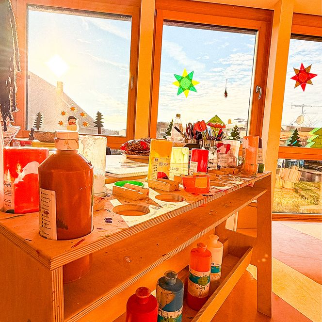 Bunte Farbtöpfe, Pinsel und Bastelutensilien stehen auf einem Holztisch im Sonnenlicht.