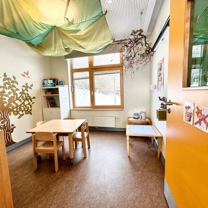 Ein Gruppenraum im Kindergarten mit Tisch und Stühlen, bunter Wandgestaltung und einem großen Fenster mit Blick nach draußen.