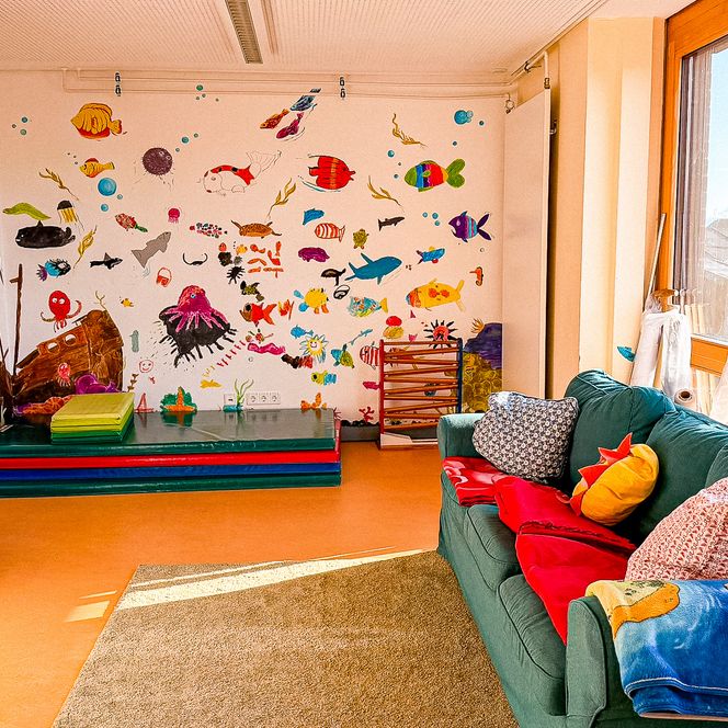 Ein heller Raum mit grüner Couch, bunten Kissen und einer Wand voller gemalter Meerestiere.