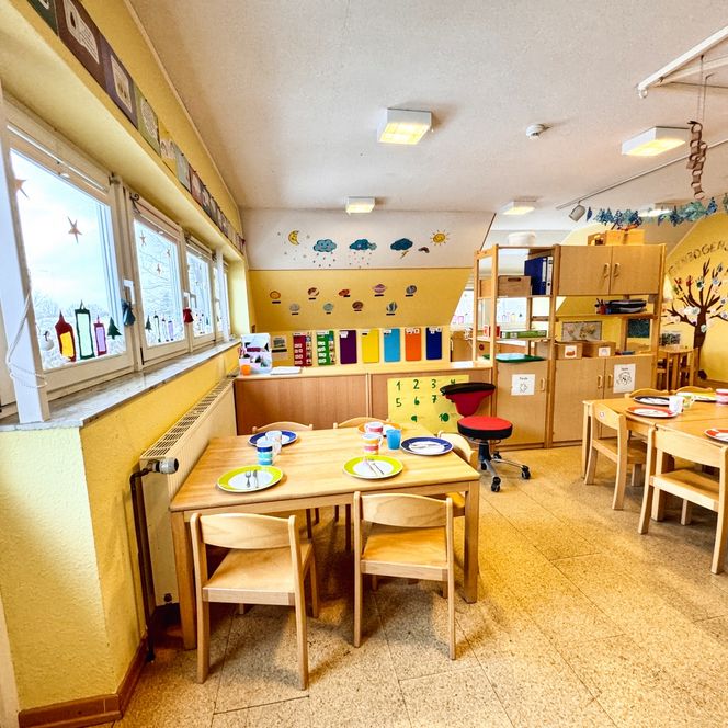 Ein heller Ess- und Gruppenraum im Kindergarten mit gedeckten Holztischen, Regalen, bunter Wandgestaltung und dekorierten Fenstern.
