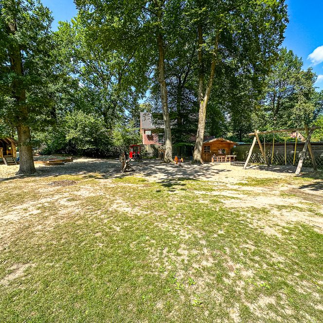 Großes Außengelände des Kindergartens Oldendorf/Luhe mit Spielhäusern, Sandflächen, Schaukelgerüst und Bäumen.