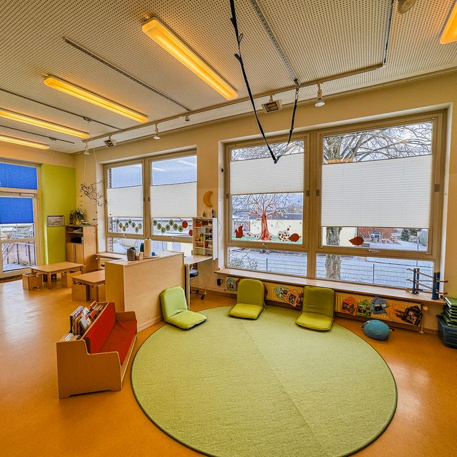 Heller Gruppenraum in der Kinderkrippe Amelinghausen mit grünem Teppichkreis, Sitzkissen, Holzmöbeln und großen Fenstern.