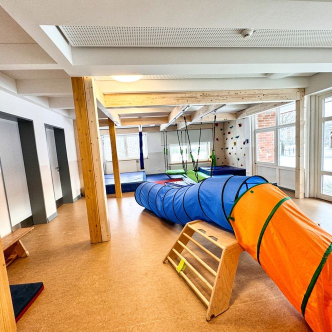 Ein heller Turn- und Spielraum im Kindergarten mit Kletterwand, Matten, Hängeschaukeln sowie einem blauen und orangefarbenen Kriechtunnel.