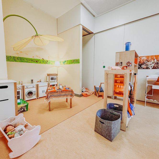 Spielbereich im Kindergarten Rehlingen mit Puppenecke, Kinderküche, Spieltisch und großem Dekoblatt an der Wand.