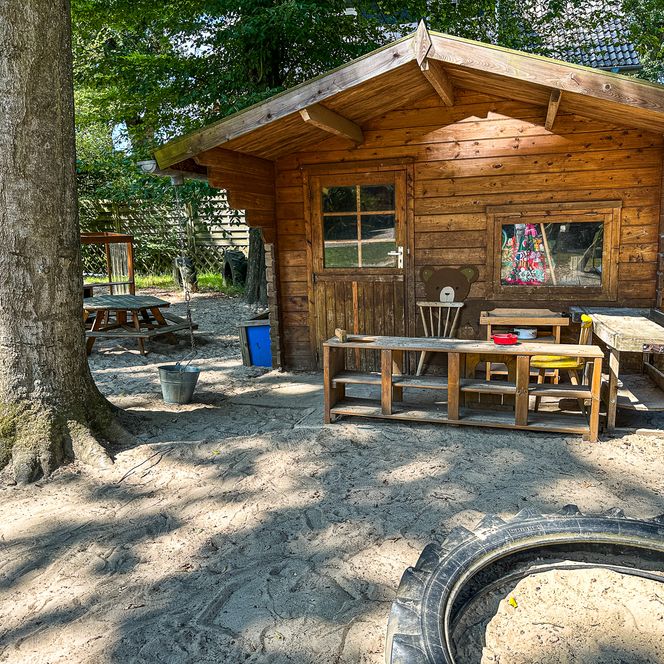 Holzspielhaus im Außengelände des Kindergartens Oldendorf/Luhe mit Sandfläche, Tisch und Spielgeräten.